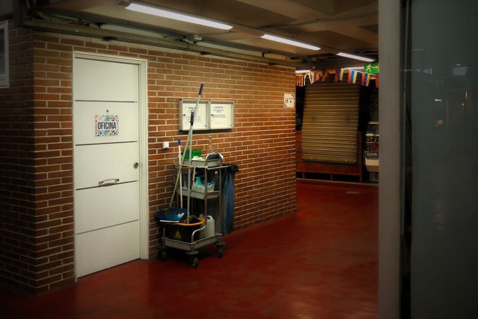 A cleaner in a plain uniform wiping down a desk in an empty open-plan office, mop bucket and supplies trolley beside them