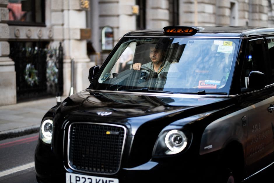 A licensed private hire car parked at a rank at dusk with the driver checking a phone at the wheel