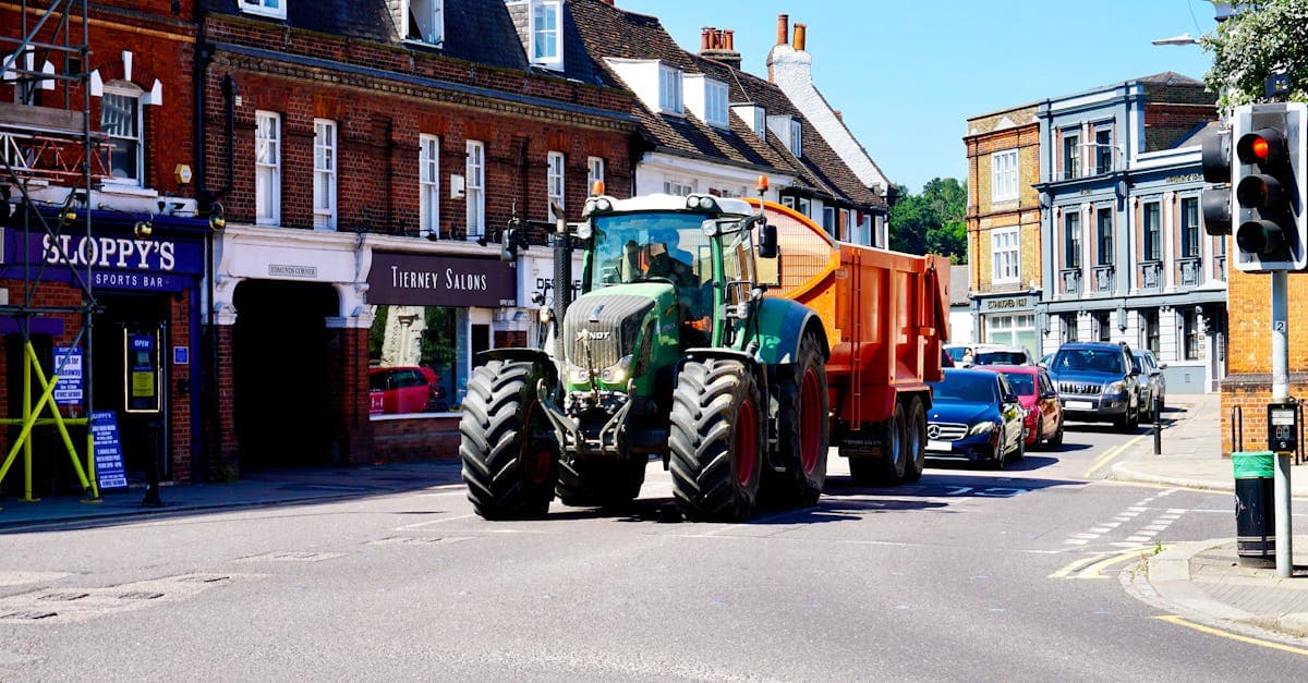 A green tractor drives through Hertford town center, surrounded by cars and shops.