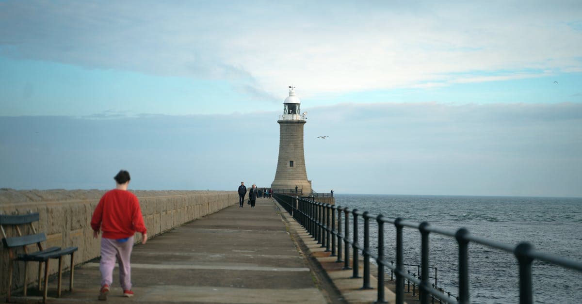 Coastal pier with a lighthouse and people walking during daytime, offering a serene seaside view.